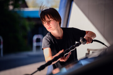 A woman intensely focused on washing her car at a self-service station. She uses a high-pressure...