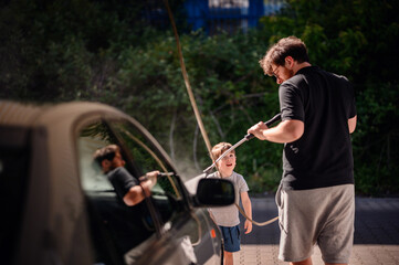A father teaching his young son to wash a car with a high-pressure hose at a self-service station....