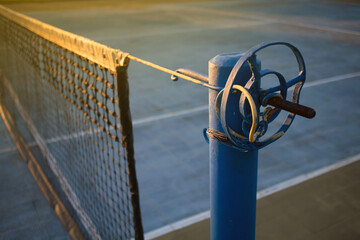 view of amateur blue tennis court at sunset, with close up of net post with rudimentary torque...
