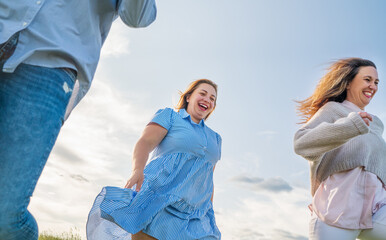 Portrait of three cheerful smiling women running by the meadow. Low angle photo shot on the blue sky with clouds. Woman's friendship, relations, and happiness concept image.