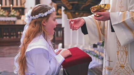 Young Caucasian Catholic Girl in White Alb Festive Outfit Receiving First Holy Communion Sacrament...
