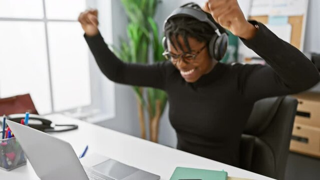 Excited woman with dreadlocks celebrates success at her office desk, wearing headphones in a professional indoor setting.