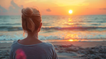 A woman with a bun hairstyle stands at the beach, gazing at the serene sunset over the ocean.