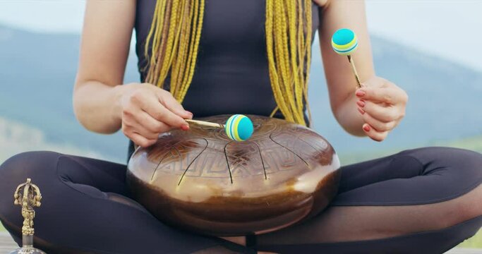 Anonymous woman with dreadlocks playing on glucophone, sitting in lotus position outdoors. Close up of red haired female making rhythm on tongue drum, while meditating. Concept of yoga, music.