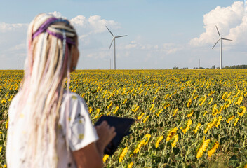 young woman with a tablet in field with sunflowers, wind turbines for green energy production,...
