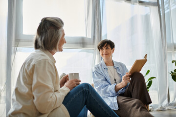 Two women, a loving mature lesbian couple, sitting on the floor in front of a window.