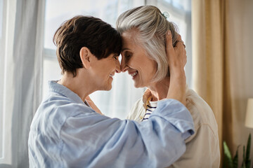 Two older women embrace lovingly in front of a window.