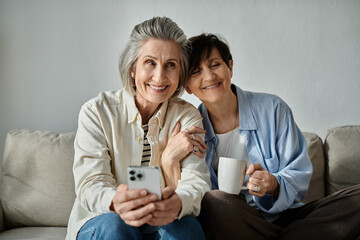 Two women, a loving mature lesbian couple, sit on a couch, engrossed in a cell phone.