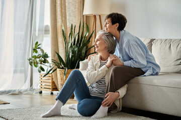 Two women, a loving mature lesbian couple, sit side by side on a cozy couch in a bright living room.