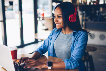 Cheerful female black model resting in coffee shop listening favorite music in headphones and...