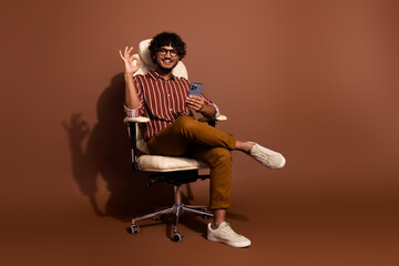 Full body portrait of nice young man sit chair show okay symbol wear striped shirt isolated on...