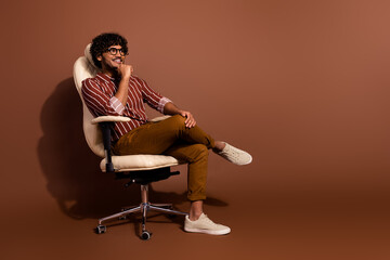 Fototapeta premium Full body portrait of nice young man sit chair look empty space wear striped shirt isolated on brown color background