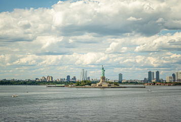 Naklejka premium New York, NY, USA - August 1, 2023: Statue of Liberty on its island in wide landscape showing New Jersey coastline under blue cloudscape