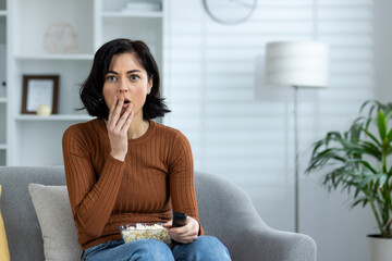 Woman watching a horror movie at home, eating popcorn and looking shocked