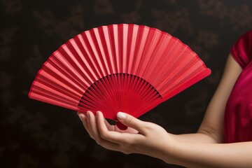 Close-up on delicate hands presenting a vibrant red folding fan against a dark backdrop - Powered by Adobe