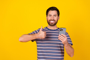 Photo of satisfied man with beard dressed striped t-shirt hold smartphone showing thumb up isolated on vibrant yellow color background