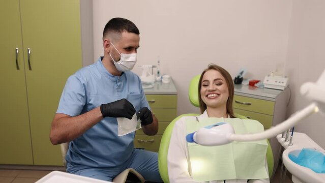 Dental chair, Oral hygiene, Dental examination. Woman is seated in dental chair while dentist in blue scrubs uses mouth retractor to perform examination.