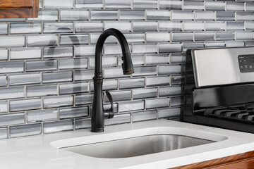 A kitchen faucet detail with a grey glass subway tile backsplash, rubbed bronze faucet, and a white...