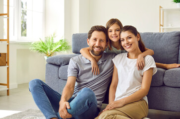 Cheerful smiling happy family sitting on the floor in the living room at home with their child girl and looking at camera. Cute daughter hugging her parents on sofa. Love and family leisure concept.