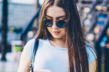 Stylish female skater with long brunette hair and sunglasses holding srateboard in hand while...