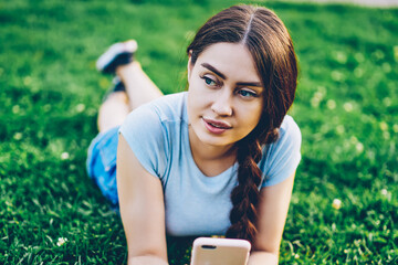 Pondering beautiful young woman with telephone in hands lying on green lawn while browsing internet websites.Attractive hipster girl with braid resting on grass while checking mail on smartphone