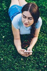 Brunette attractive young woman with braid resting on green grass in park enjoying summer weekend and sending notification to friend on smartphone device via high speed internet connection