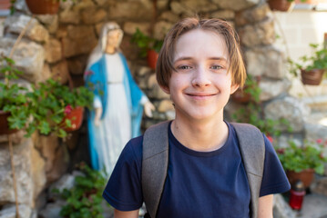 An 11-year-old smiling Catholic boy with a backpack against the background of a statue of the Virgin Mary.