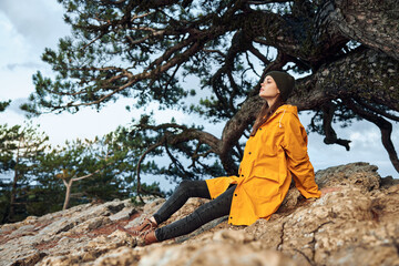 Naklejka premium A woman in a yellow coat enjoying the peaceful serenity of nature on top of a rocky cliff near a pine tree