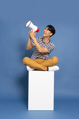 Young Asian man wearing striped shirt shout loudly and holding megaphone and sitting on white box...