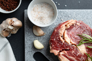 Top view of fresh raw beef steak garnished with rosemary on cutting board. Scattered salt and garlic cloves on kitchen counter. Close-up