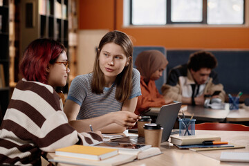 Young Caucasian and Asian girls sitting at table in university library doing homework or preparing for tests
