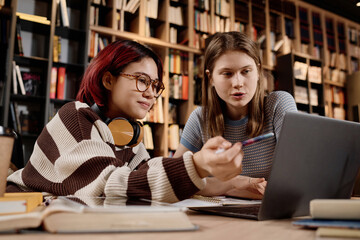 Medium shot of two girls sitting at table looking at laptop screen and discussing issue while...