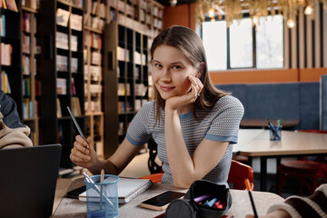 Medium portrait of modern Caucasian girl holding pencil sitting at table in university or college...