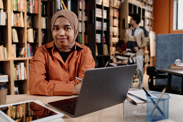 Modern Muslim girl wearing hijab sitting at desk in college library studying with use of laptop,...