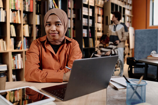 Modern Muslim girl wearing hijab sitting at desk in college library studying with use of laptop, copy space