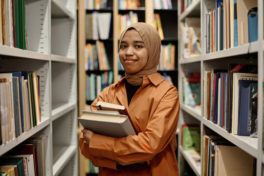Medium portrait of joyful Muslim girl wearing hijab holding books standing in college library smiling at camera, copy space