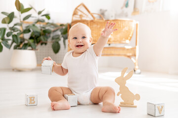 Fototapeta premium a little baby girl of six months with blue eyes in a white bodysuit plays on the floor of the children's room with wooden toys cubes smiles and rejoices, early development of the child