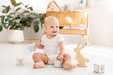 a little baby girl of six months with blue eyes in a white bodysuit plays on the floor of the...