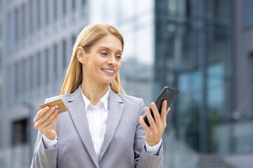 A confident businesswoman in a grey suit holds a credit card and smartphone while standing in a...