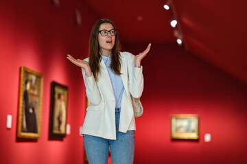 Thoughtful young Caucasian woman wearing glasses and looking at exhibition. Concept of Museum Day.