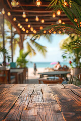 A wooden table in the foreground with a blurred background of a beachside cafe. The background shows sandy beach views through large windows, casual seating, tropical plants, and customers