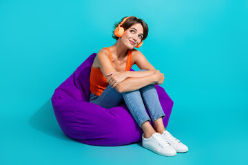 Full size photo of gorgeous good mood girl sit on bean bag in headphones look at promo empty space isolated on blue color background