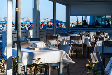 A photorealistic depiction of an outdoor restaurant with white tables and chairs arranged under large parasols on a sandy beach. The turquoise ocean gently laps at the shore in the background