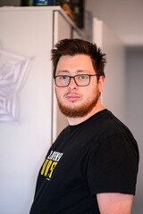 Portrait of a man with glasses and a beard standing indoors, looking at the camera with a neutral expression.