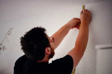 A man with glasses and a beard uses a tape measure to measure the height on a wall for an...