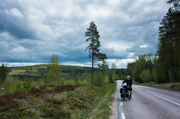 Cyclist with touring bicycle is walking up the road in nordic forest in overcast weather, Long distance cyclist solo travel in Sweden
