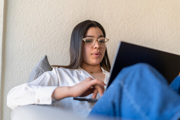 A Latina woman in glasses sits on a couch, intently working on her laptop in a bright, serene...