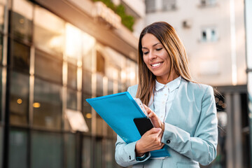 A smiling female office worker with a folder with files and a mobile phone in her hands is walking on the way to work.