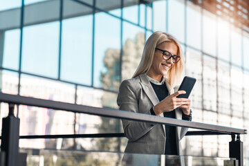 Smiling businesswoman with mobile phone standing on the balcony of an office building. Happy female...