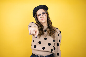 Young beautiful brunette woman wearing french beret and glasses over yellow background pointing...